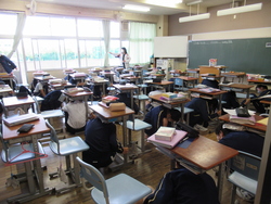 地震の避難訓練の放送を聞いて、机の下にもぐる生徒たちの写真